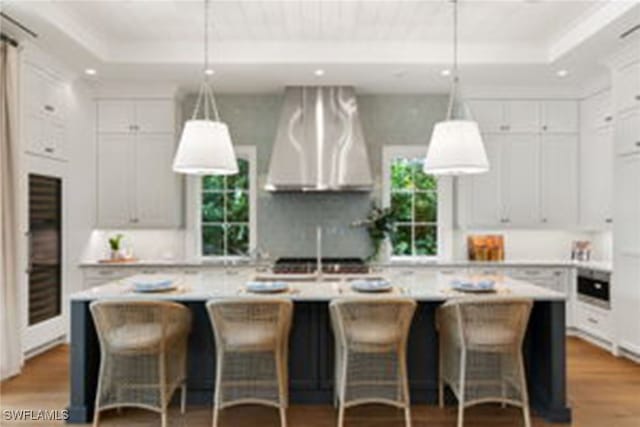 kitchen with a tray ceiling, hanging light fixtures, wall chimney exhaust hood, and an island with sink