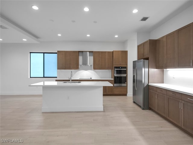 kitchen with stainless steel appliances, a center island with sink, wall chimney exhaust hood, sink, and light hardwood / wood-style flooring