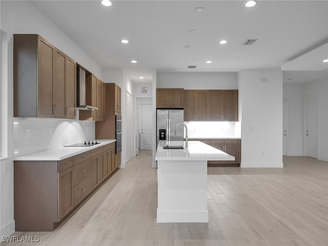 kitchen with stainless steel appliances, light hardwood / wood-style floors, a center island with sink, sink, and wall chimney range hood