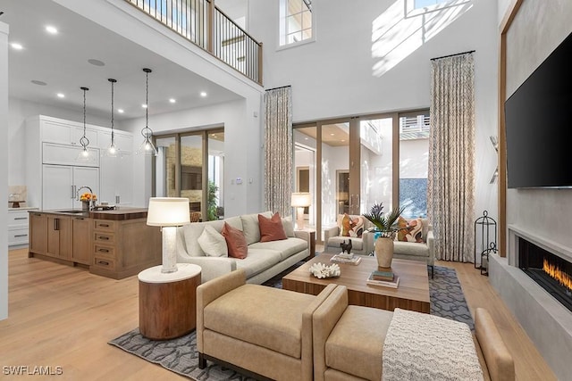 living room with light wood-type flooring, a towering ceiling, plenty of natural light, a warm lit fireplace, and recessed lighting