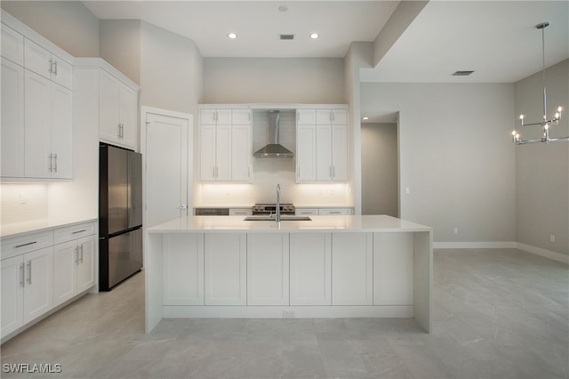 kitchen with stainless steel refrigerator, pendant lighting, white cabinets, wall chimney exhaust hood, and a kitchen island with sink