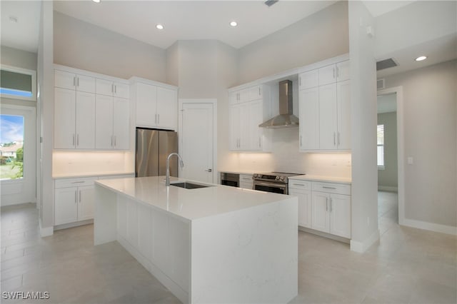 kitchen with a center island with sink, white cabinetry, wall chimney range hood, appliances with stainless steel finishes, and sink