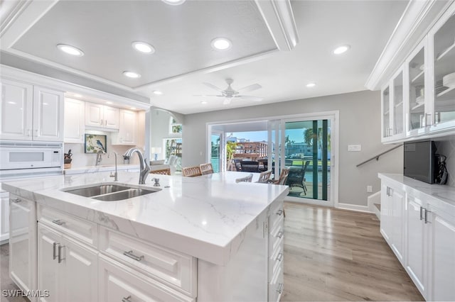 kitchen featuring white microwave, a sink, white cabinetry, a center island with sink, and glass insert cabinets