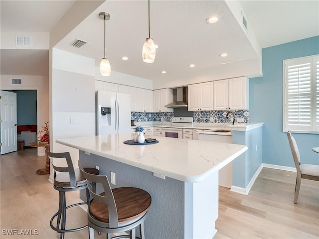 kitchen featuring white appliances, white cabinets, wall chimney range hood, decorative backsplash, and decorative light fixtures