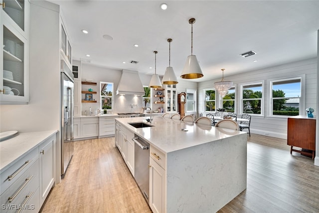 kitchen with sink, an island with sink, custom range hood, pendant lighting, and appliances with stainless steel finishes