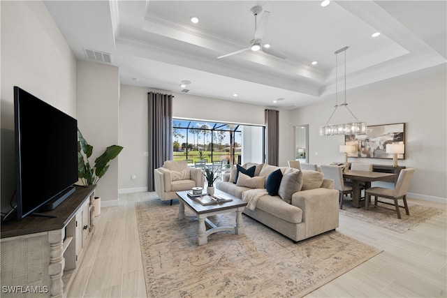 living room featuring a tray ceiling, crown molding, ceiling fan with notable chandelier, and light wood-type flooring