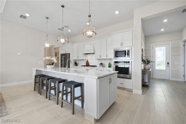 kitchen with appliances with stainless steel finishes, premium range hood, a kitchen island with sink, white cabinetry, and hanging light fixtures