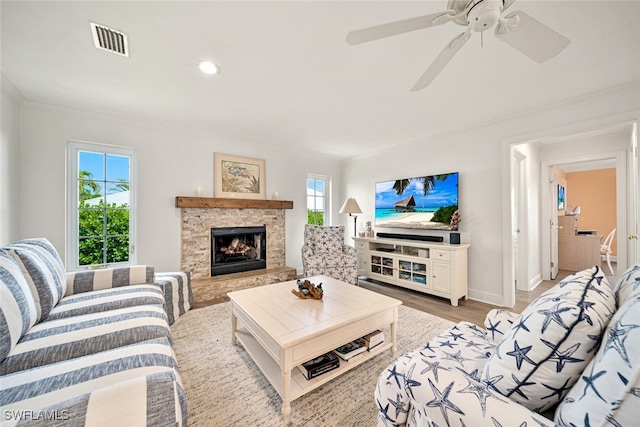 living room with ceiling fan, a stone fireplace, light wood-type flooring, and ornamental molding