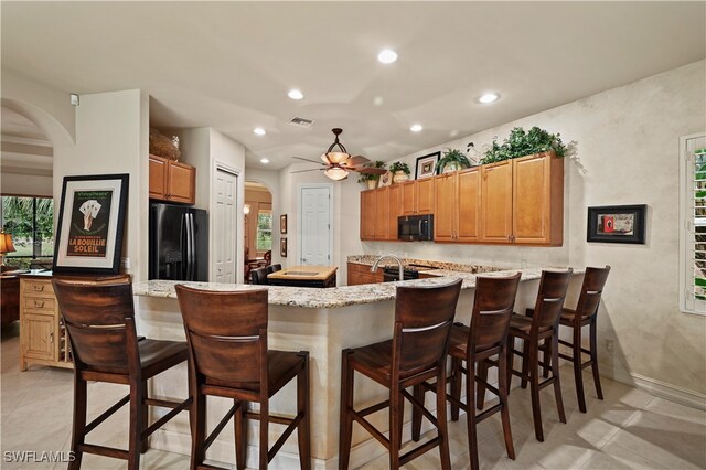 kitchen featuring light stone counters, ceiling fan, black appliances, light tile patterned floors, and a breakfast bar area