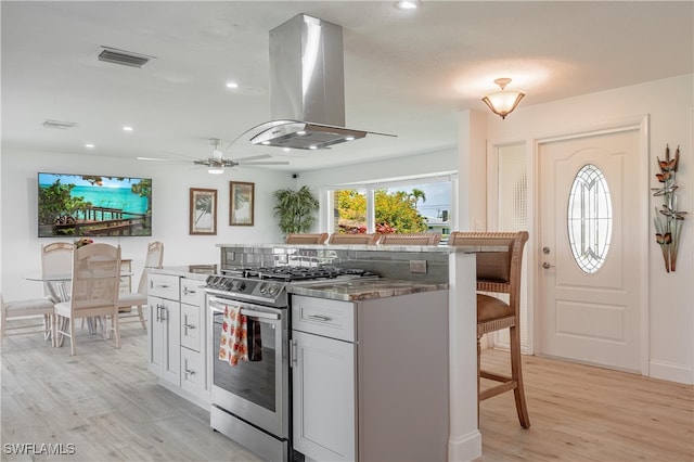 kitchen with ceiling fan, stainless steel gas stove, light hardwood / wood-style flooring, island exhaust hood, and a kitchen bar