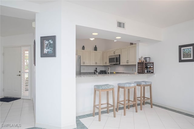 kitchen featuring cream cabinets, appliances with stainless steel finishes, a kitchen breakfast bar, light tile patterned floors, and decorative backsplash