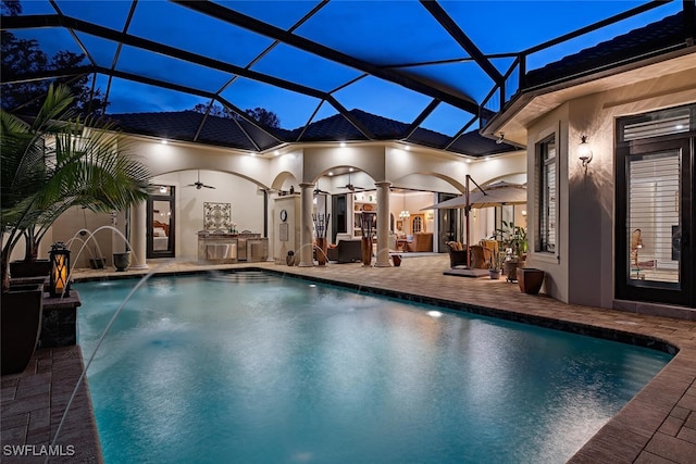 pool at dusk with a patio, pool water feature, glass enclosure, and ceiling fan