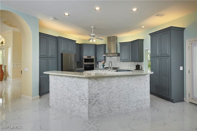 kitchen with wall chimney exhaust hood, ceiling fan, backsplash, a kitchen island with sink, and appliances with stainless steel finishes