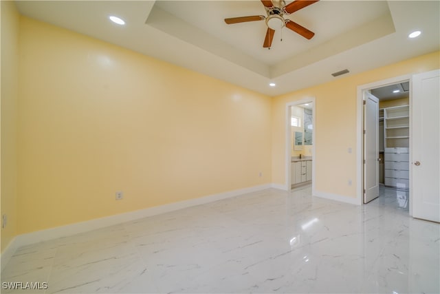 unfurnished bedroom with ceiling fan, ensuite bath, a spacious closet, and a tray ceiling