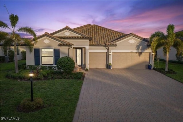 mediterranean / spanish-style house with a yard, an attached garage, stucco siding, a tile roof, and decorative driveway