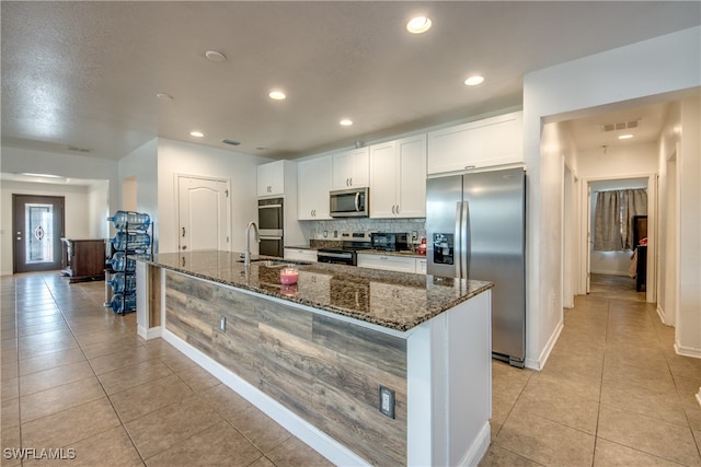 kitchen with white cabinets, appliances with stainless steel finishes, a kitchen island with sink, and dark stone counters