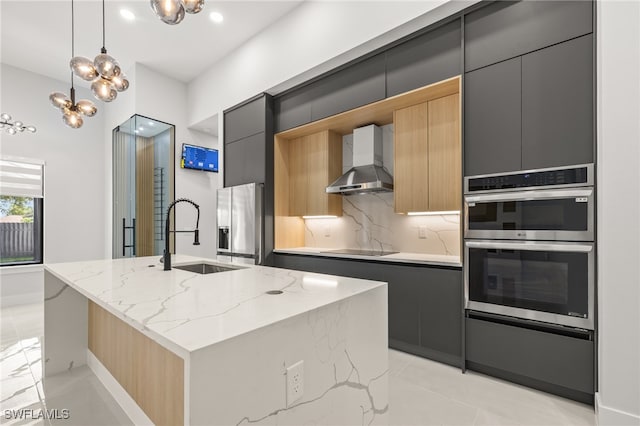 kitchen with stainless steel appliances, a center island with sink, light stone counters, wall chimney exhaust hood, and pendant lighting