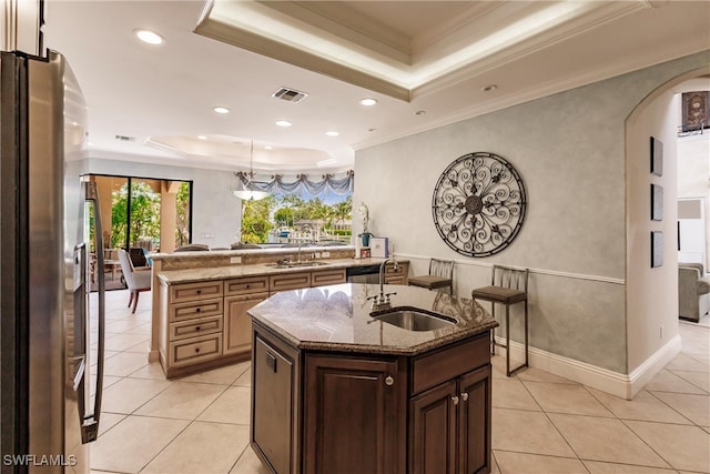 kitchen featuring light tile patterned flooring, stainless steel refrigerator, a raised ceiling, light stone countertops, and an island with sink