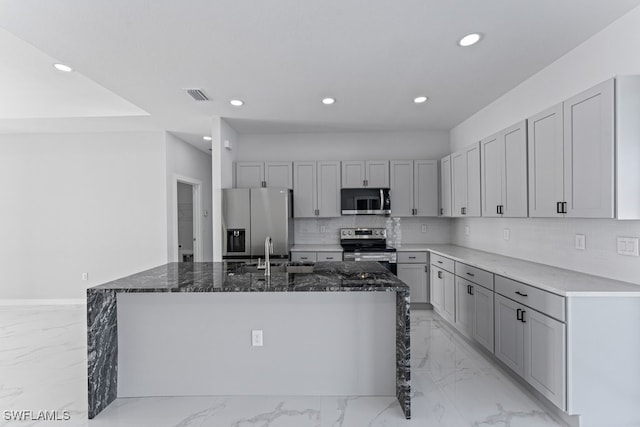 kitchen with backsplash, dark stone counters, sink, an island with sink, and stainless steel appliances