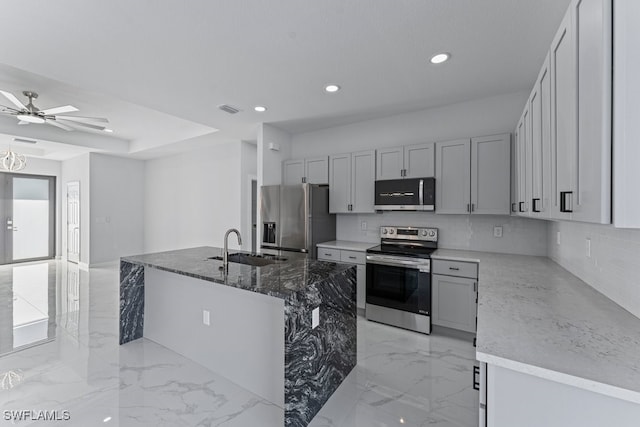 kitchen with sink, ceiling fan, dark stone countertops, an island with sink, and appliances with stainless steel finishes