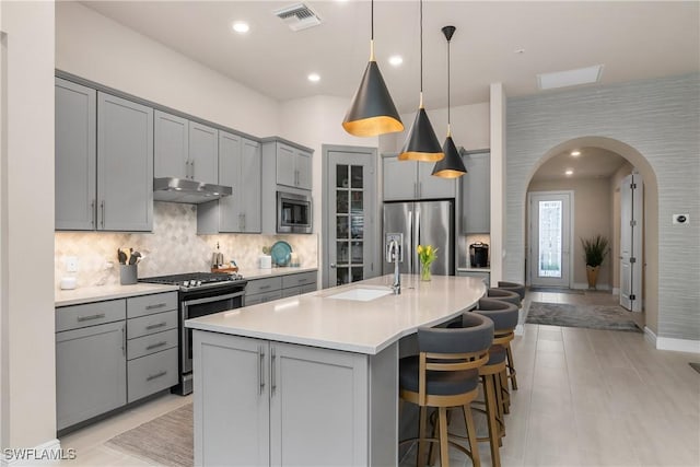 kitchen with backsplash, decorative light fixtures, gray cabinets, a center island with sink, and appliances with stainless steel finishes