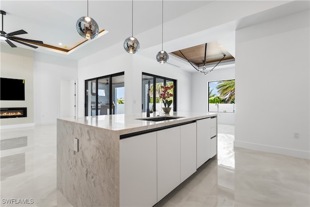 kitchen with light stone countertops, a raised ceiling, an island with sink, pendant lighting, and white cabinets