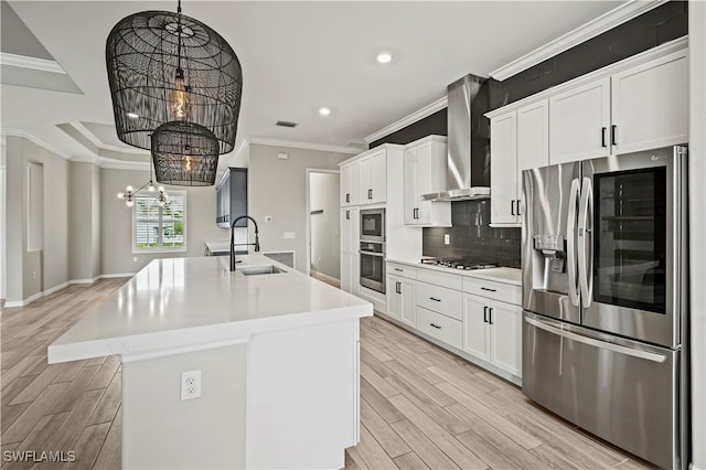 kitchen featuring white cabinetry, stainless steel appliances, wall chimney range hood, a chandelier, and a center island with sink