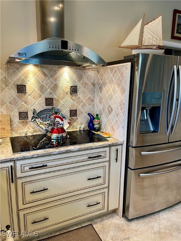 kitchen with stainless steel fridge, light stone counters, wall chimney range hood, cream cabinetry, and light tile patterned flooring