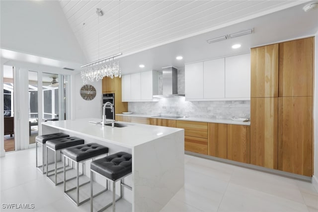 kitchen with decorative backsplash, black electric stovetop, wall chimney exhaust hood, a large island with sink, and white cabinets