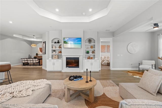 living room featuring built in shelves, hardwood / wood-style flooring, a raised ceiling, and ceiling fan