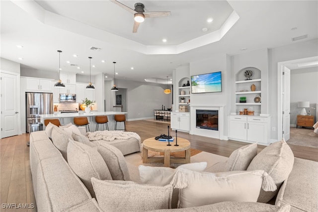 living room with built in shelves, light wood-type flooring, ceiling fan, and a raised ceiling