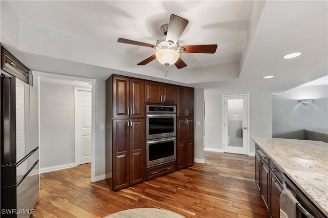 kitchen with ceiling fan, dark wood-type flooring, stainless steel appliances, light stone counters, and a tray ceiling