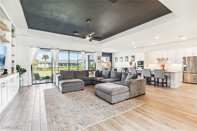 living room with a raised ceiling, ceiling fan, and light wood-type flooring