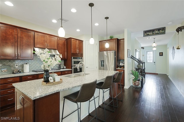 kitchen featuring dark wood-type flooring, decorative backsplash, an island with sink, appliances with stainless steel finishes, and decorative light fixtures