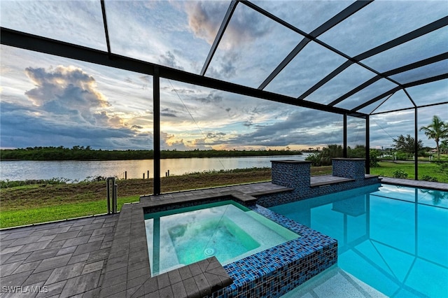 pool at dusk featuring a water view, an in ground hot tub, glass enclosure, a patio, and a yard