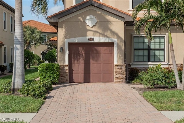 exterior space featuring decorative driveway, stone siding, an attached garage, and stucco siding