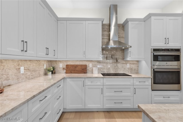 kitchen with wall chimney exhaust hood, black electric cooktop, double oven, tasteful backsplash, and light stone counters