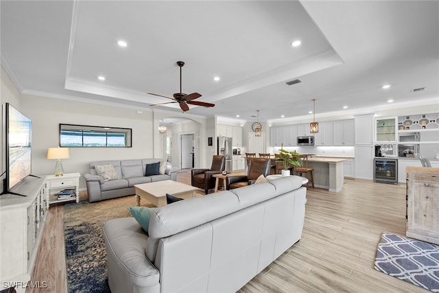 living room with ceiling fan, beverage cooler, crown molding, light hardwood / wood-style floors, and a tray ceiling