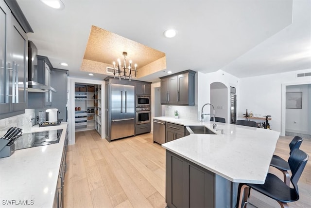 kitchen featuring built in appliances, wall chimney exhaust hood, sink, a raised ceiling, and pendant lighting