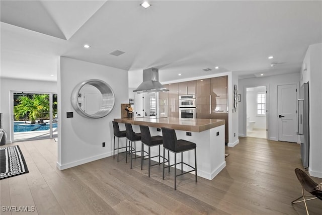 kitchen with light hardwood / wood-style flooring, appliances with stainless steel finishes, a kitchen breakfast bar, kitchen peninsula, and island exhaust hood