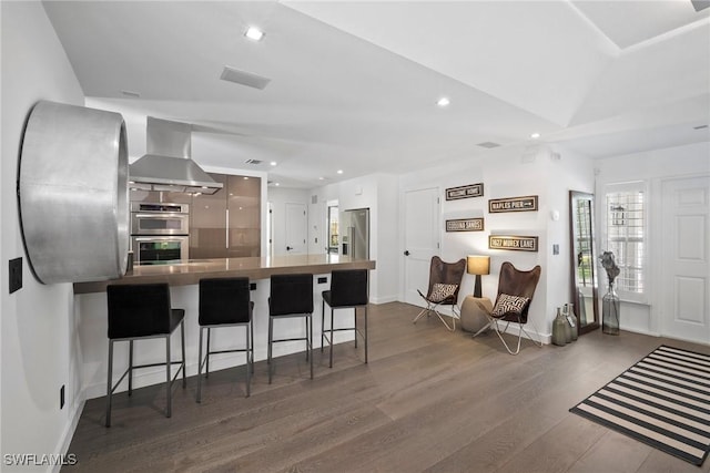 kitchen featuring dark wood-type flooring, a kitchen breakfast bar, wall chimney range hood, kitchen peninsula, and stainless steel double oven