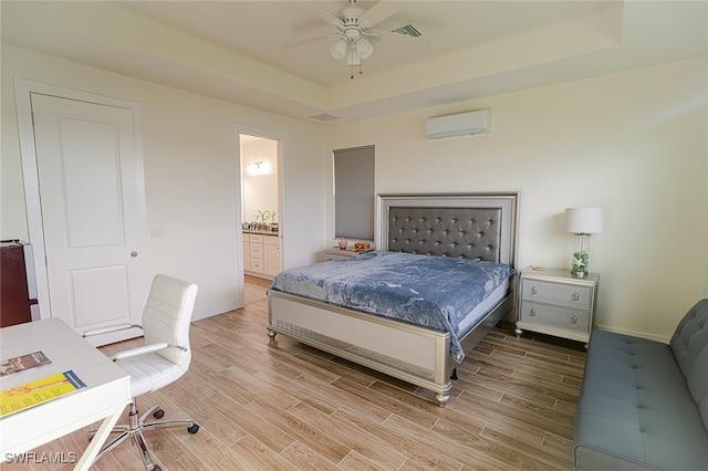 bedroom featuring a wall unit AC, light hardwood / wood-style floors, a tray ceiling, ceiling fan, and ensuite bathroom