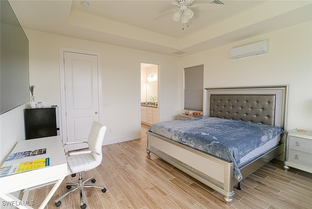 bedroom featuring ensuite bath, ceiling fan, a tray ceiling, light hardwood / wood-style flooring, and a wall mounted air conditioner