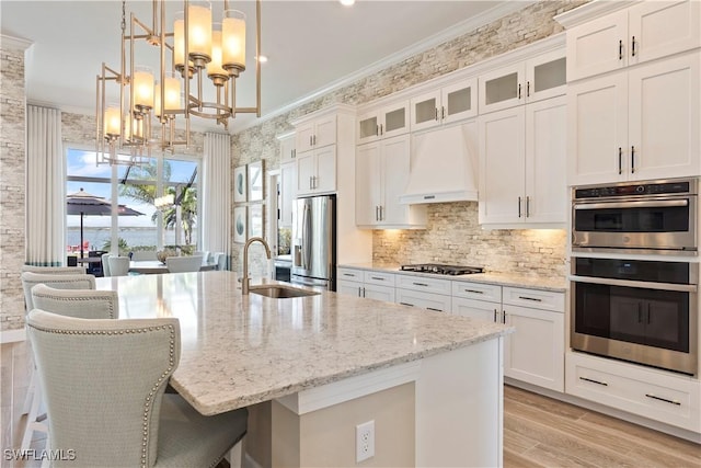 kitchen with premium range hood, appliances with stainless steel finishes, hanging light fixtures, ornamental molding, and a center island with sink