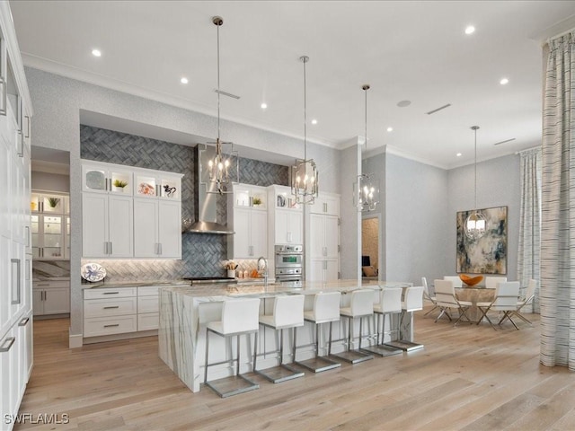 kitchen featuring wall chimney range hood, a large island with sink, glass insert cabinets, and white cabinetry