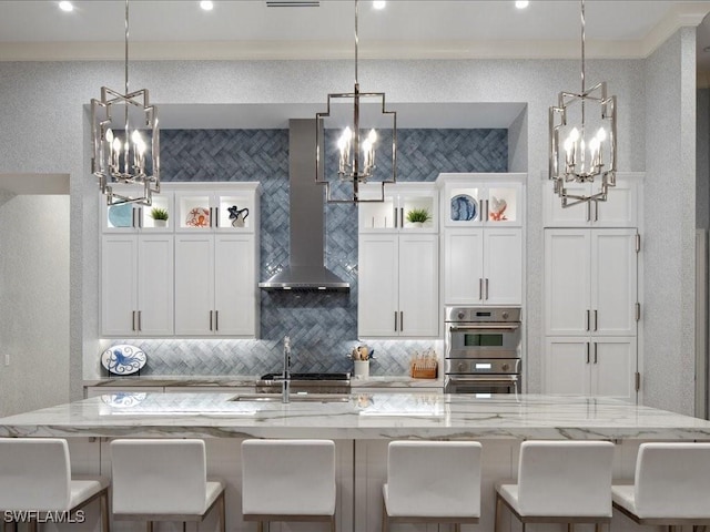 kitchen featuring wall chimney exhaust hood, glass insert cabinets, light stone countertops, pendant lighting, and a notable chandelier