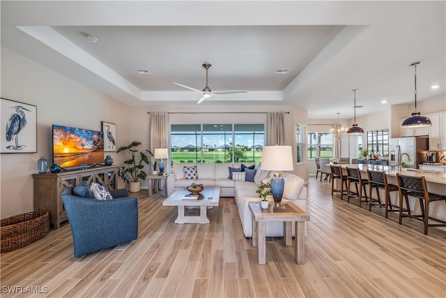 living room with ceiling fan with notable chandelier, light hardwood / wood-style flooring, and a tray ceiling