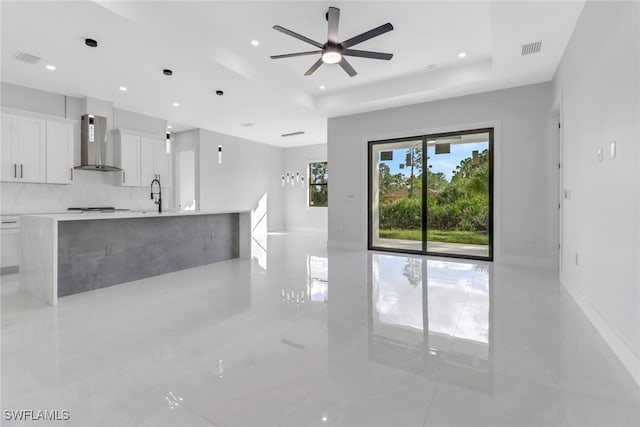 kitchen with sink, ceiling fan, a raised ceiling, white cabinets, and wall chimney range hood