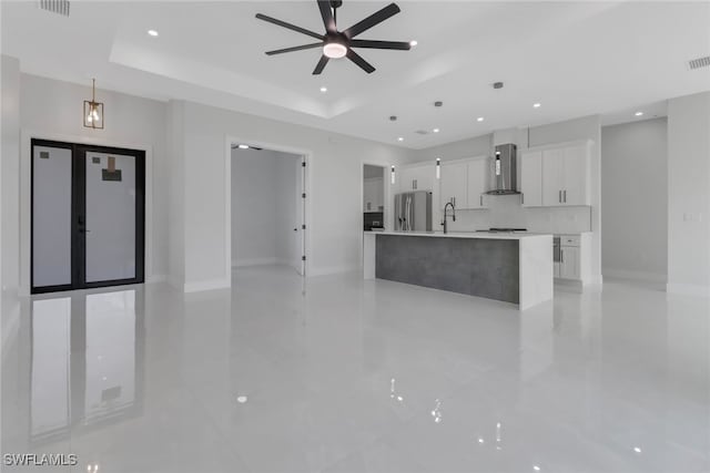 kitchen with a kitchen island with sink, a raised ceiling, wall chimney range hood, stainless steel refrigerator, and white cabinetry