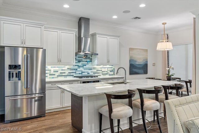 kitchen with sink, wall chimney exhaust hood, stainless steel appliances, an island with sink, and white cabinets